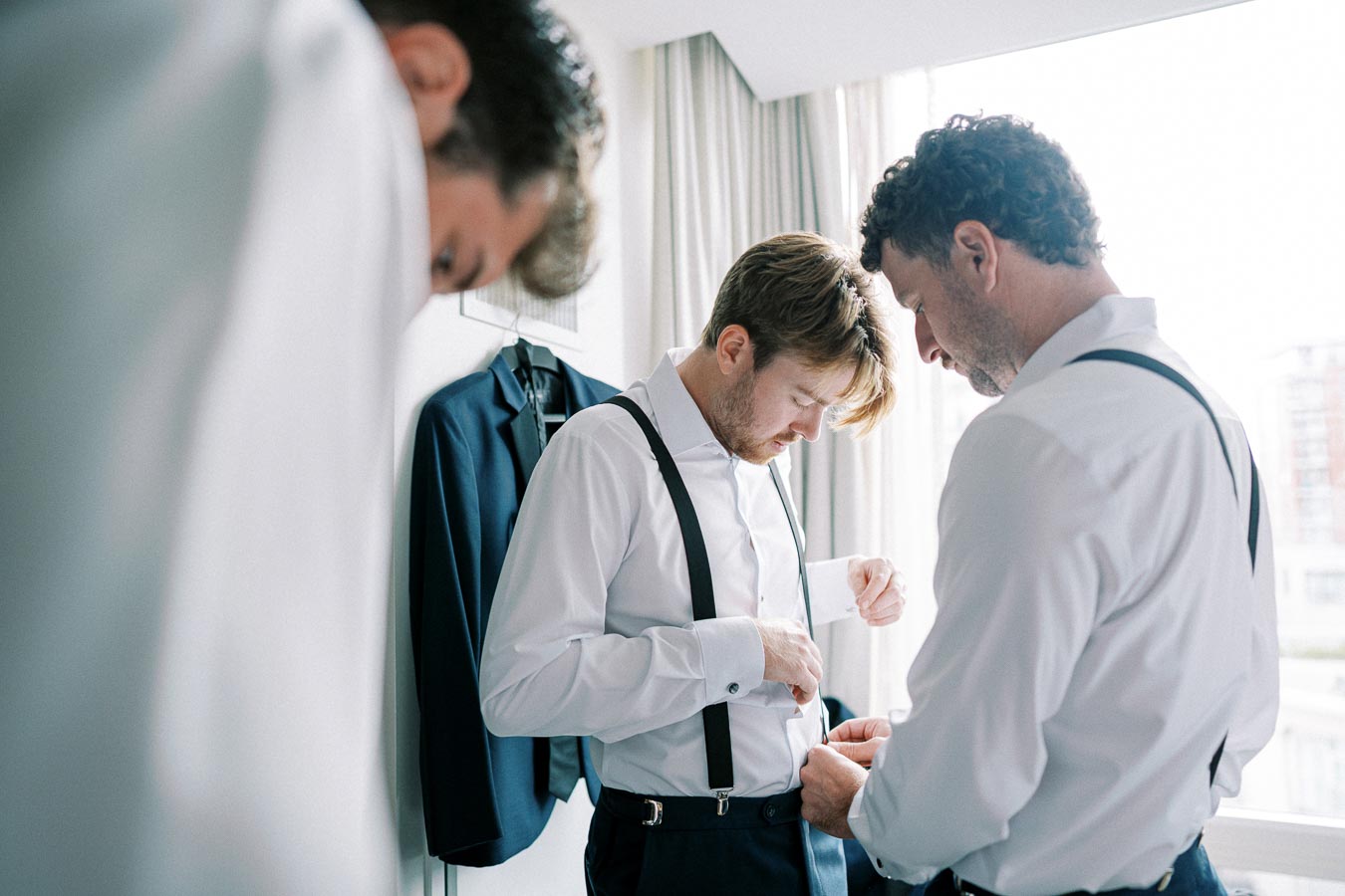 Groomsmen getting ready in a bright room, helping each other with suspenders and formal suits, showcasing wedding preparation.