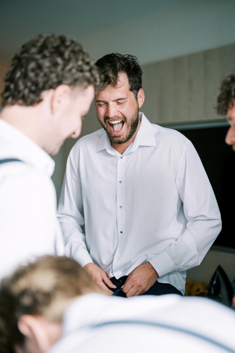 Groom laughing with groomsmen while getting ready for a wedding, wearing a white dress shirt in a joyful moment.