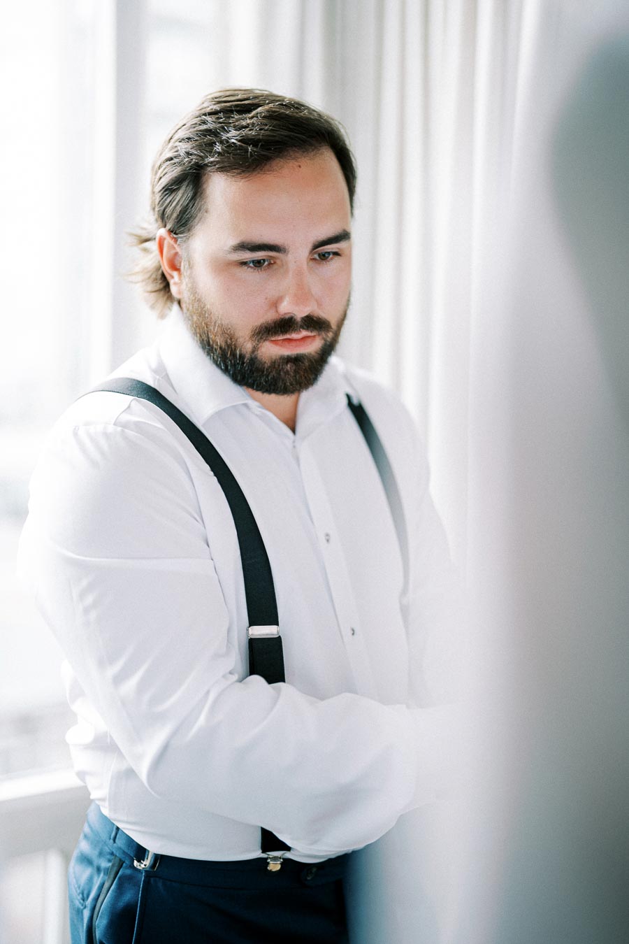 Adult man with a beard wearing a white dress shirt and dark suspenders in a softly lit room, gazing thoughtfully.