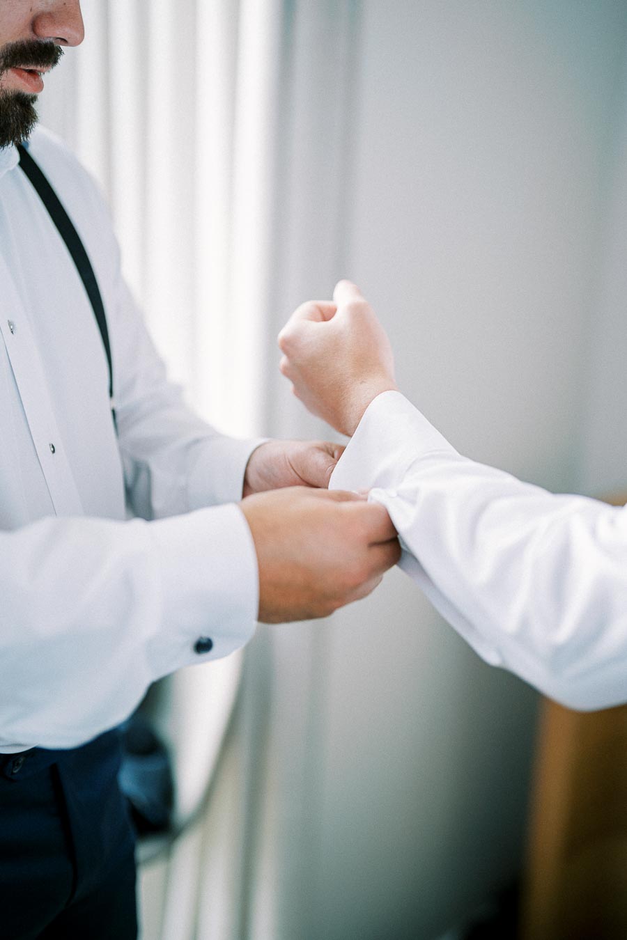 A man in a white dress shirt adjusts his cufflinks, preparing for a formal occasion, with a second person assisting him.