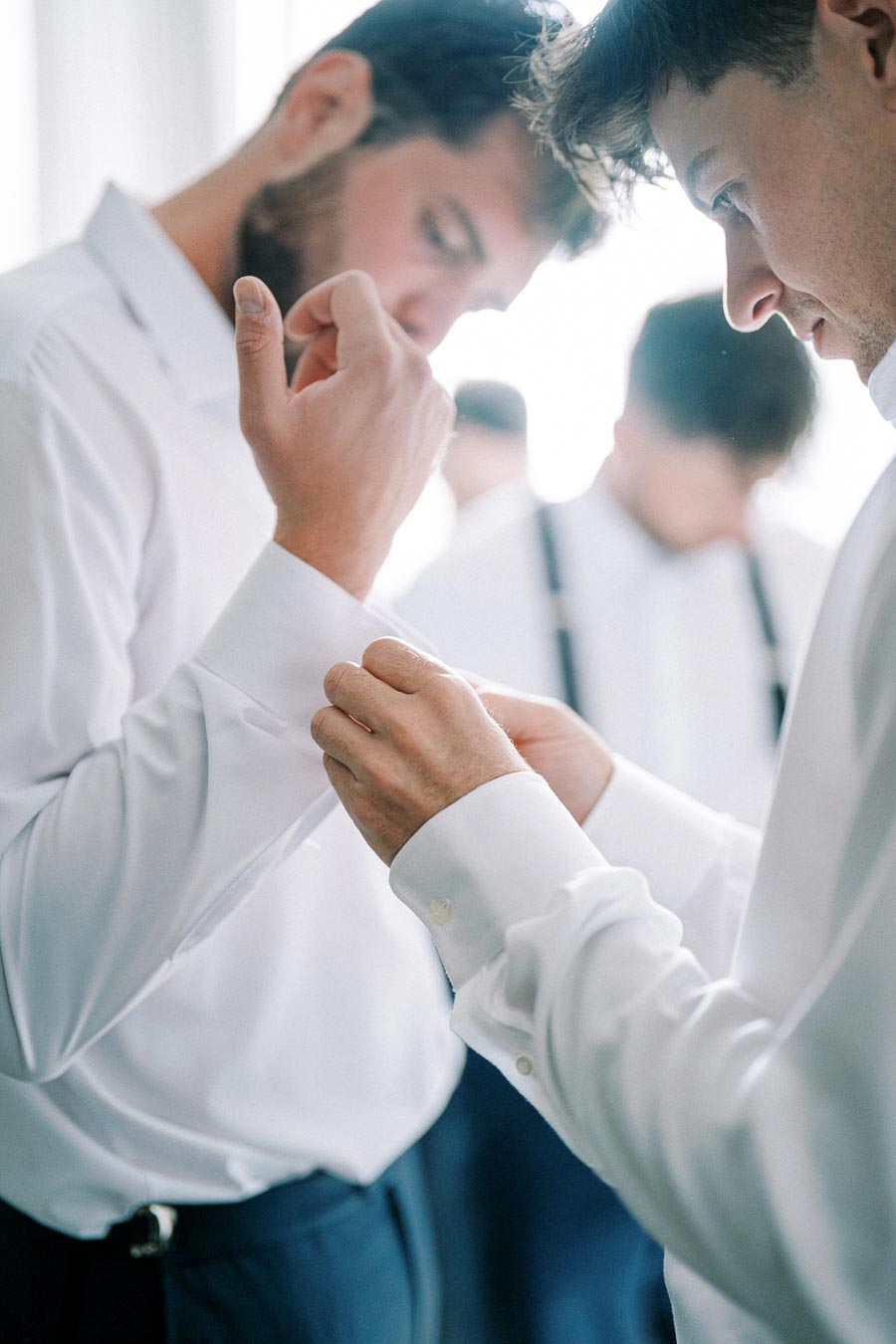 Groomsmen preparing for a wedding, adjusting cufflinks on white dress shirts in a bright room.