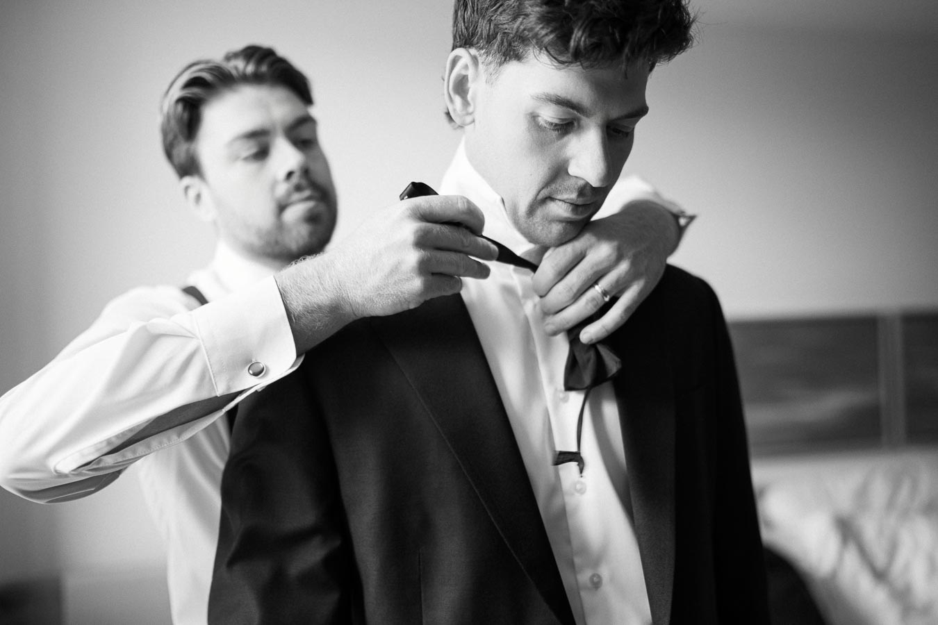 Black and white image of a groom getting help with his necktie by a groomsman before a wedding.