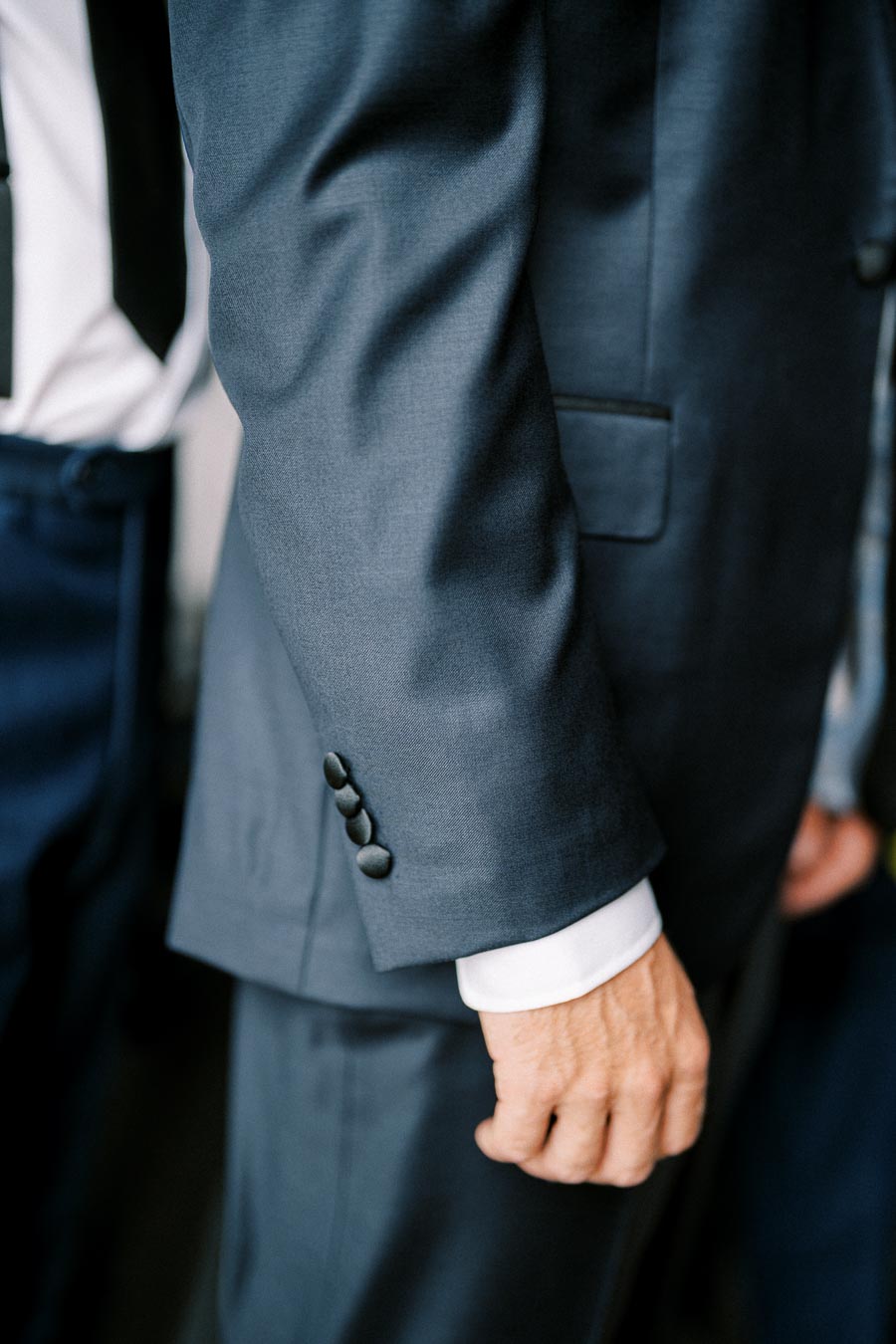Close-up of a man in a tailored navy blue suit, focusing on the sleeve buttons and white cuff, highlighting elegant menswear fashion at a formal event.