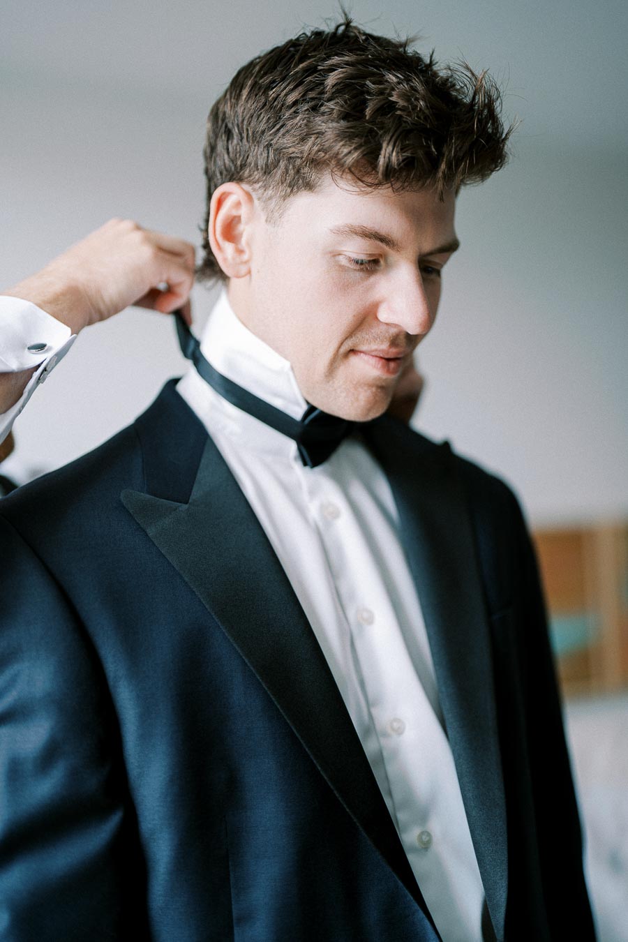 Man in formal attire being assisted with bow tie adjustment, wearing a black tuxedo jacket and white dress shirt, preparing for a formal event.