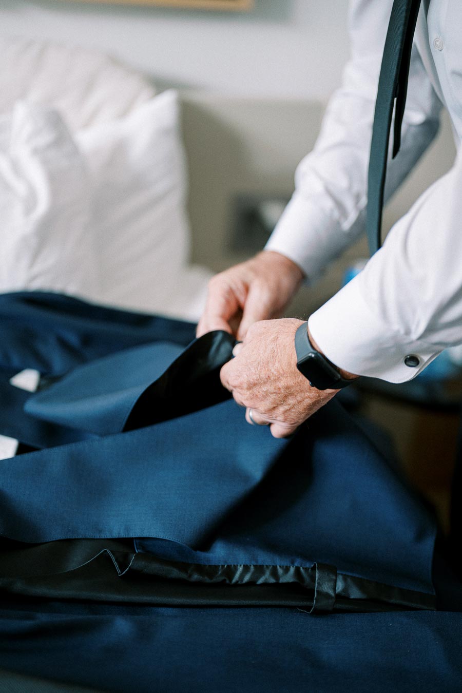 Man in formal attire holding a blue suit jacket on a bed, preparing for a formal event.