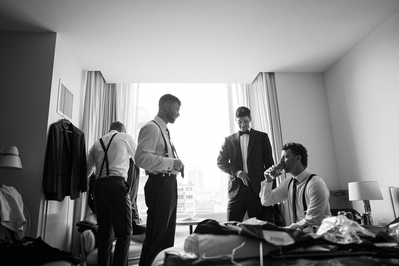Groom and groomsmen in a hotel room getting ready for a wedding, dressed in formal attire with suspenders and suits; black and white photography capturing candid pre-wedding moments.
