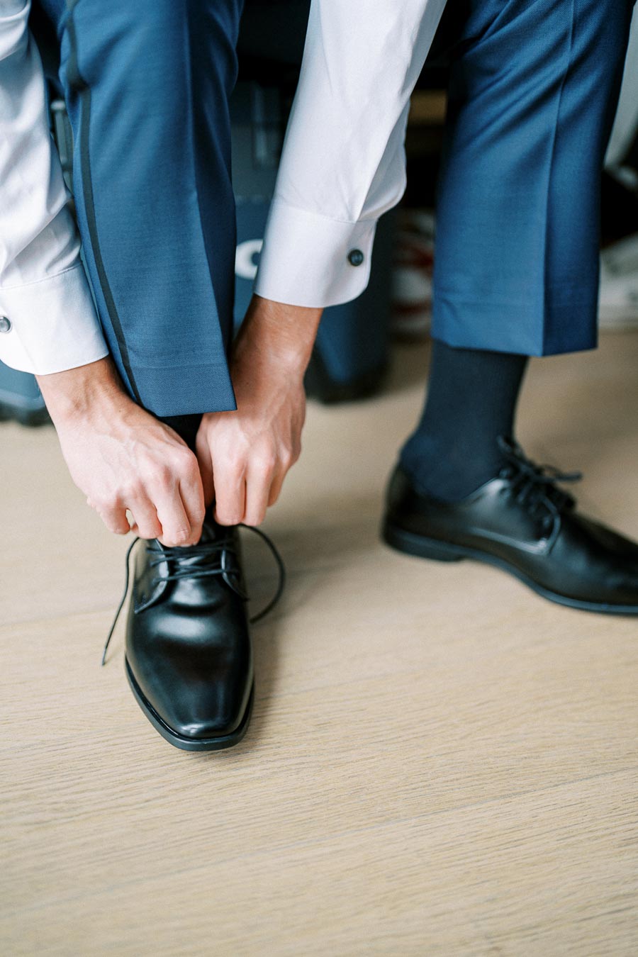 Man wearing blue suit pants and white shirt tying black leather dress shoes on a wooden floor.
