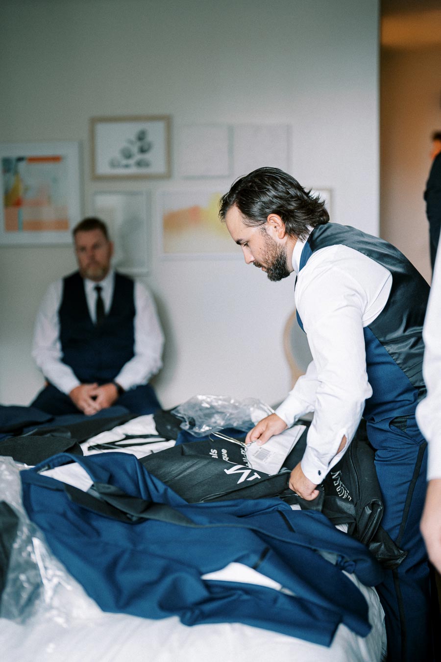 Man in formal attire organizing suits on a bed, with another man seated in the background, preparing for a special event in a well-decorated room.