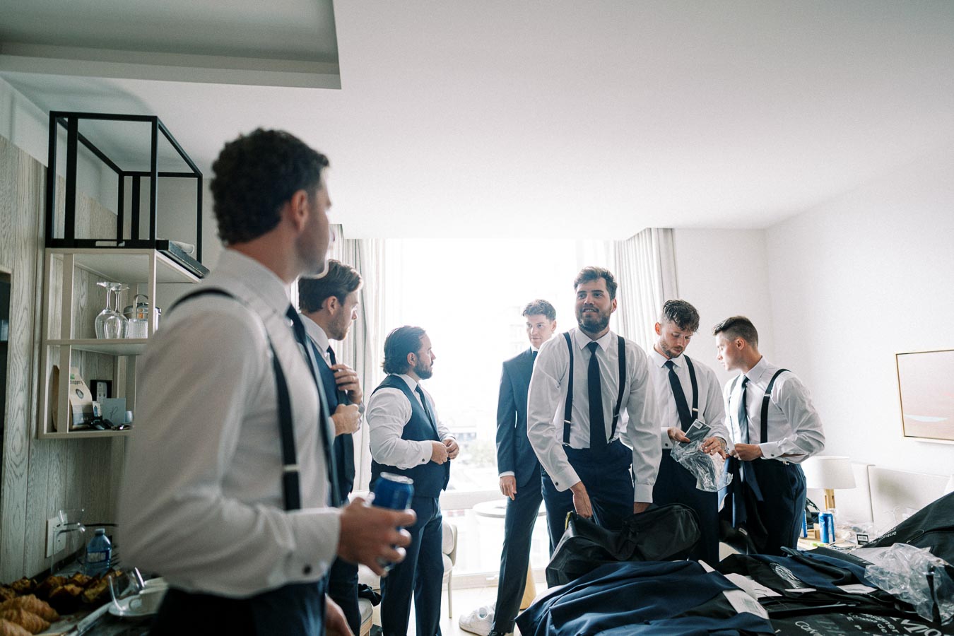Group of groomsmen in white shirts and suspenders getting ready in a bright room, preparing for a wedding ceremony.
