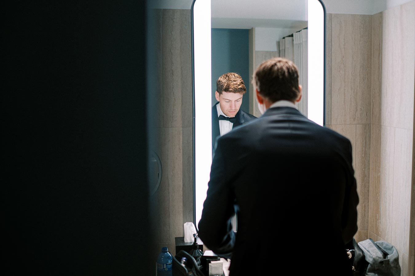 A man in a tuxedo adjusts his bow tie in front of a well-lit mirror, preparing for an elegant event in a modern bathroom.