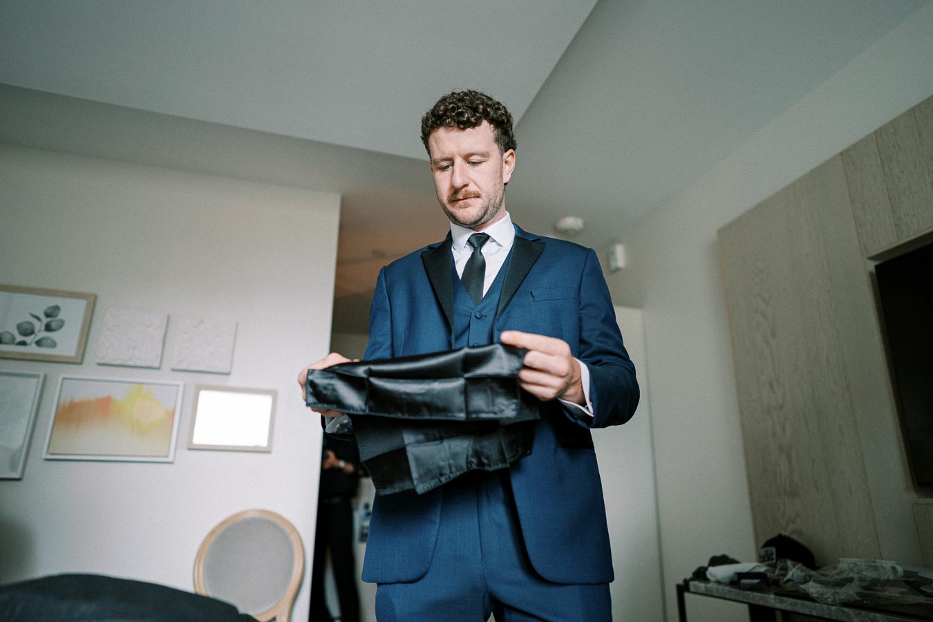A man in a blue suit standing in a modern room, holding and examining a black bow tie, preparing for a formal event.