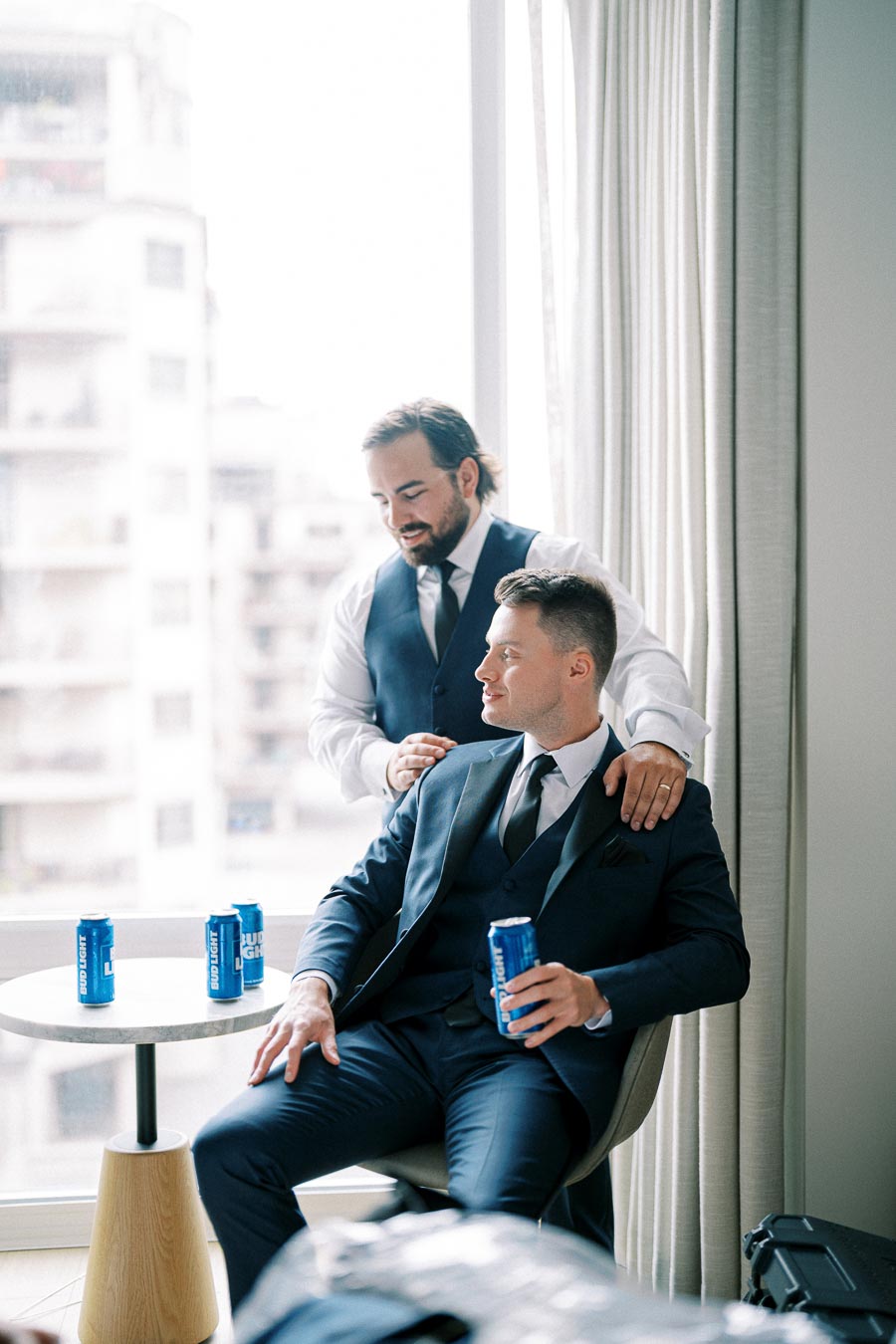 Two men in suits sharing a moment before an event, one seated holding a canned beverage, the other standing, with a cityscape view in the background.