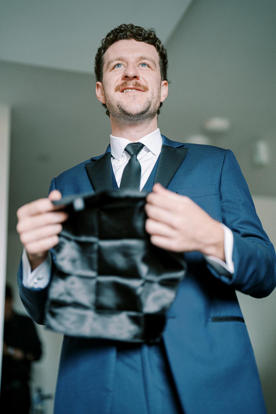 Man wearing a blue suit and black tie, smiling while holding a black cloth, standing indoors with blurred background.