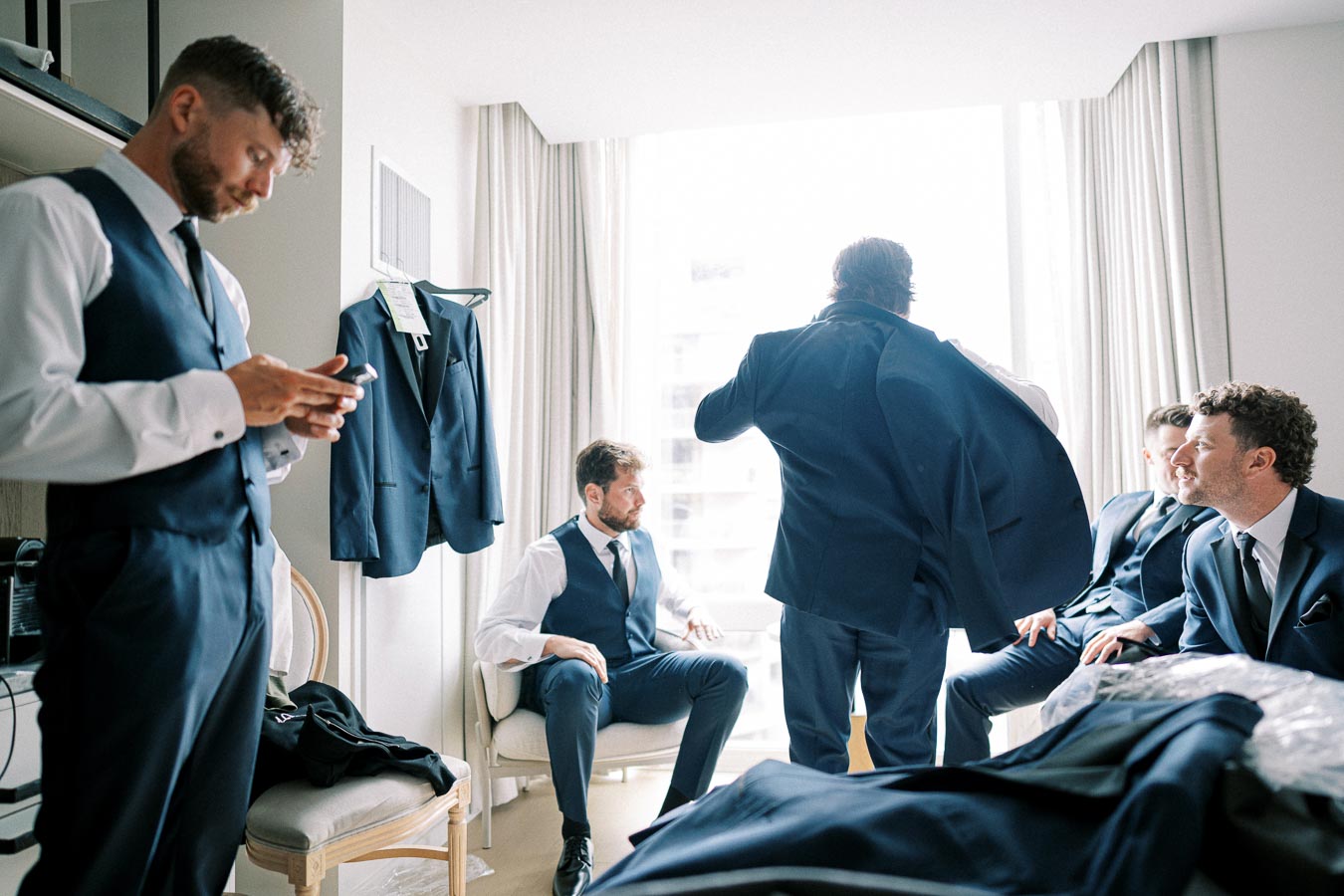 Groom and groomsmen in blue suits preparing for a wedding in a bright room, with one man checking his phone.