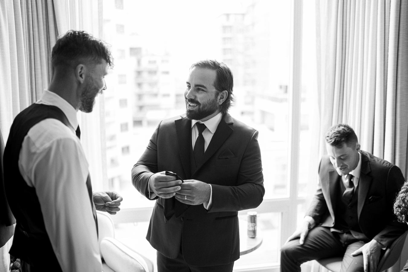 Three men preparing for a formal event, wearing suits, in a bright hotel room with large windows.