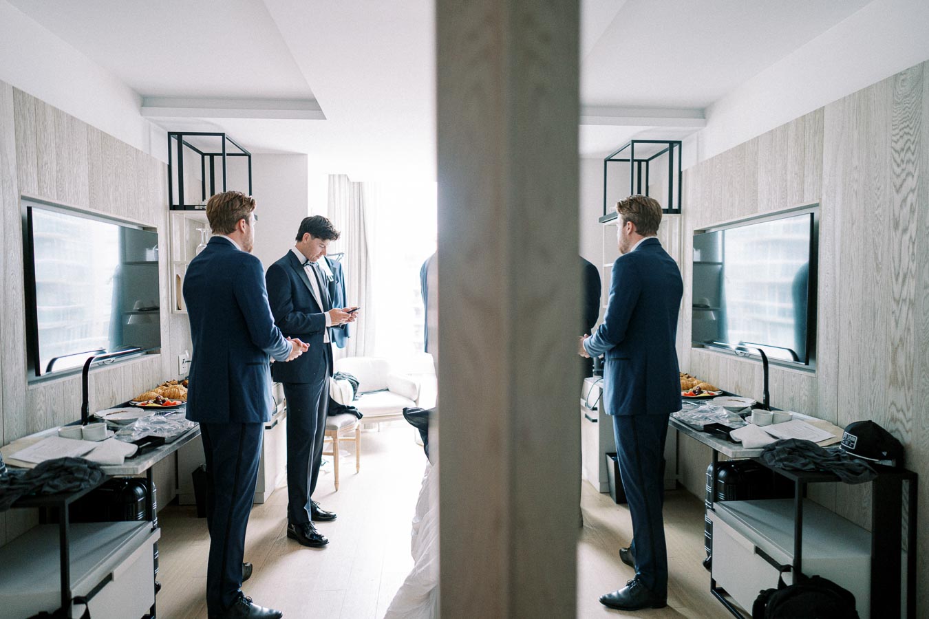 Three men in suits are gathered in a modern, well-lit room, preparing for a formal event. A central column divides the image, reflecting their busy ambiance. The room features sleek decor and a table with assorted items and snacks.