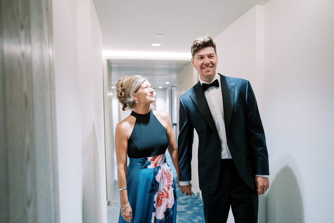 A woman in a floral gown and a man in a tuxedo smiling while walking down a hallway at an elegant event.