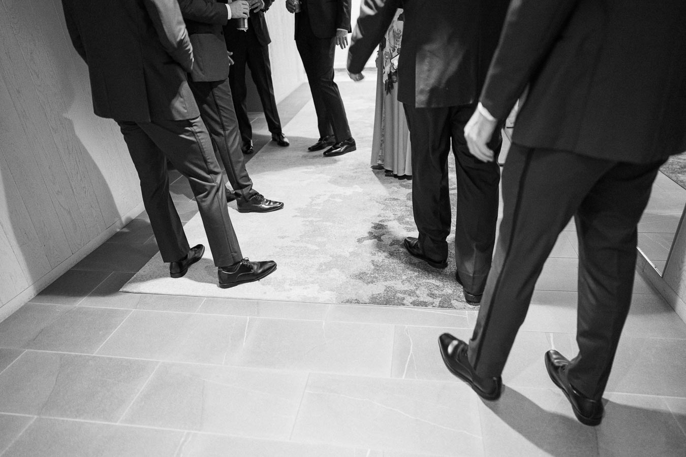 Black and white image of a group of people in formal attire, standing and conversing in a hallway.