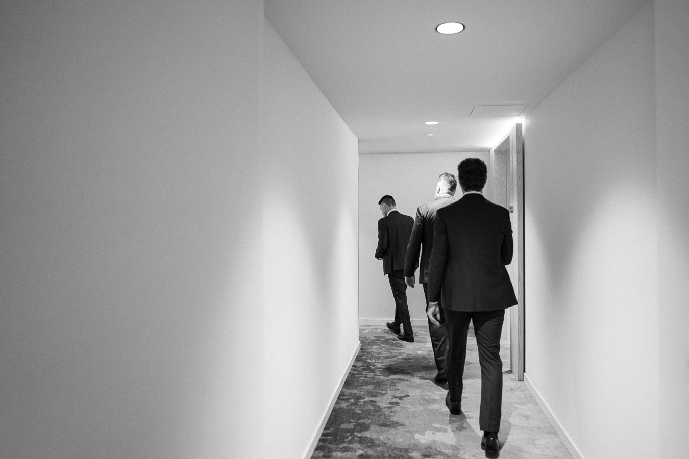 Black and white image of three businessmen in suits walking down a carpeted hallway, taken from behind; office corridor setting.