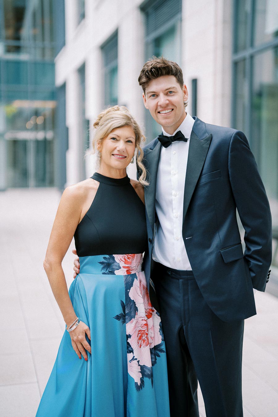 A well-dressed couple posing outdoors; the woman is wearing a blue floral gown and the man is in a black tuxedo, standing in front of a modern glass building.