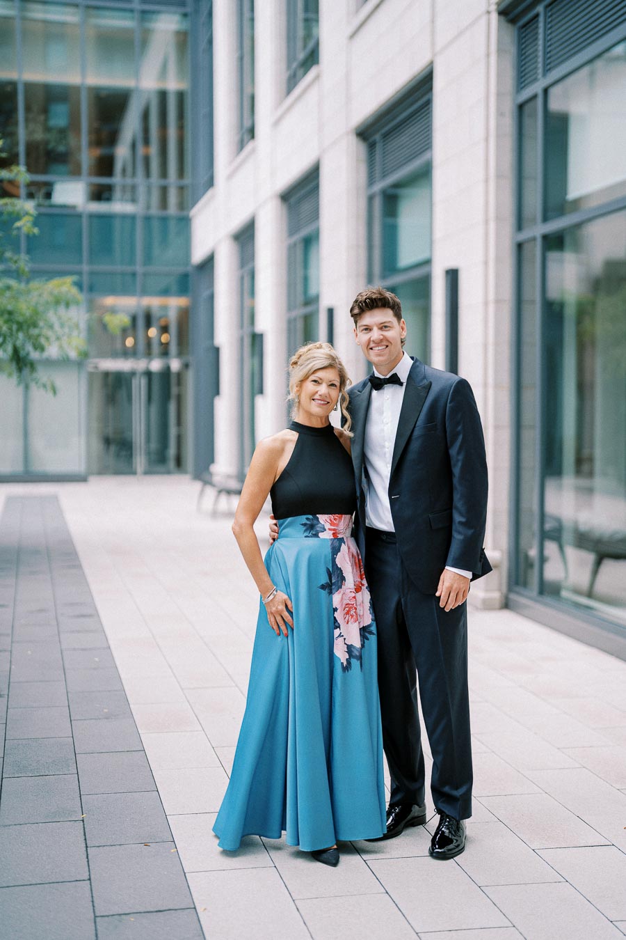 A couple in formal attire posing outdoors; the woman is wearing a blue gown with floral patterns, while the man is in a black tuxedo, against a backdrop of modern architecture.
