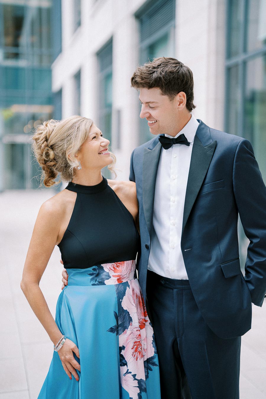 A man in a blue tuxedo and bow tie stands beside a woman in a black and floral dress, smiling at each other outdoors in an urban setting.