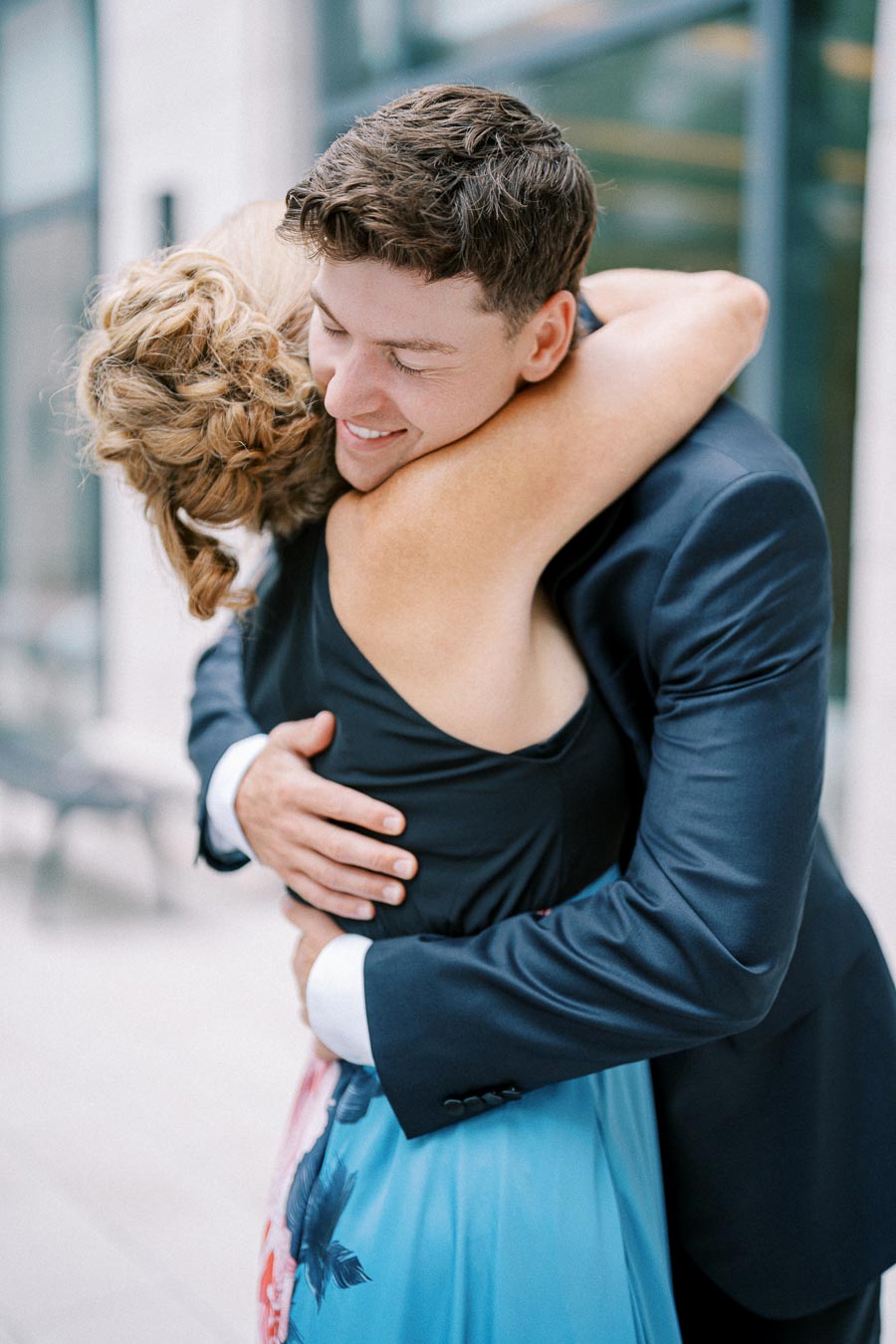 Young couple embracing warmly in formal attire, with a blurred outdoor urban setting in the background.