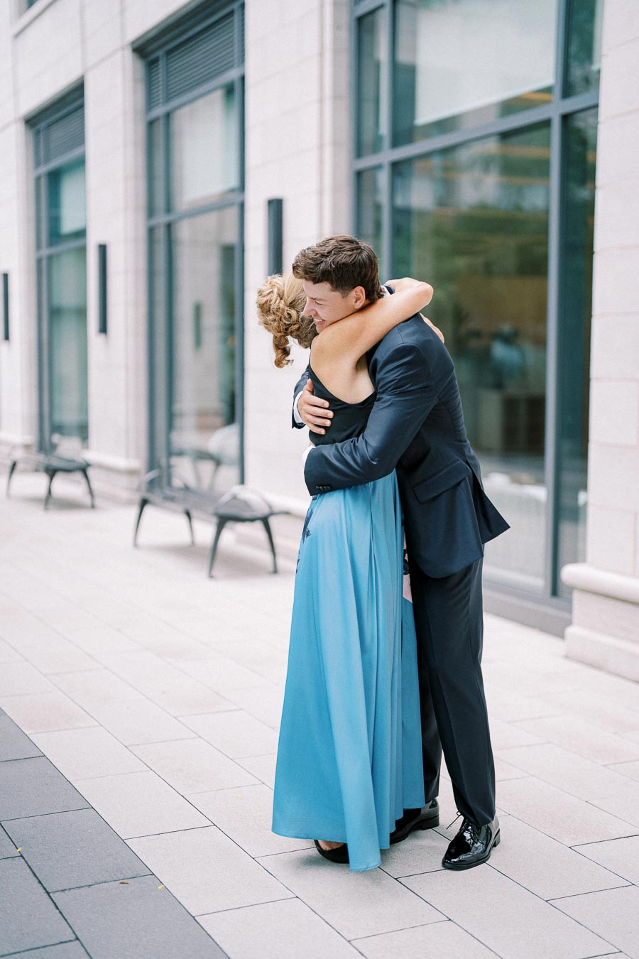 A couple embracing warmly in formal attire outside a modern building, expressing affection and happiness.