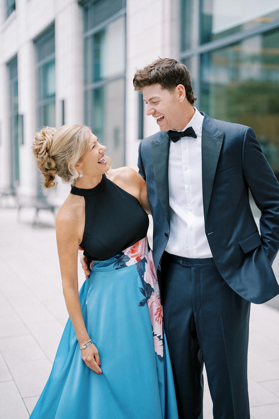 A joyful couple dressed in formal attire, with the woman in a black top and blue floral skirt and the man in a navy blue tuxedo, laughing and embracing in an urban setting.