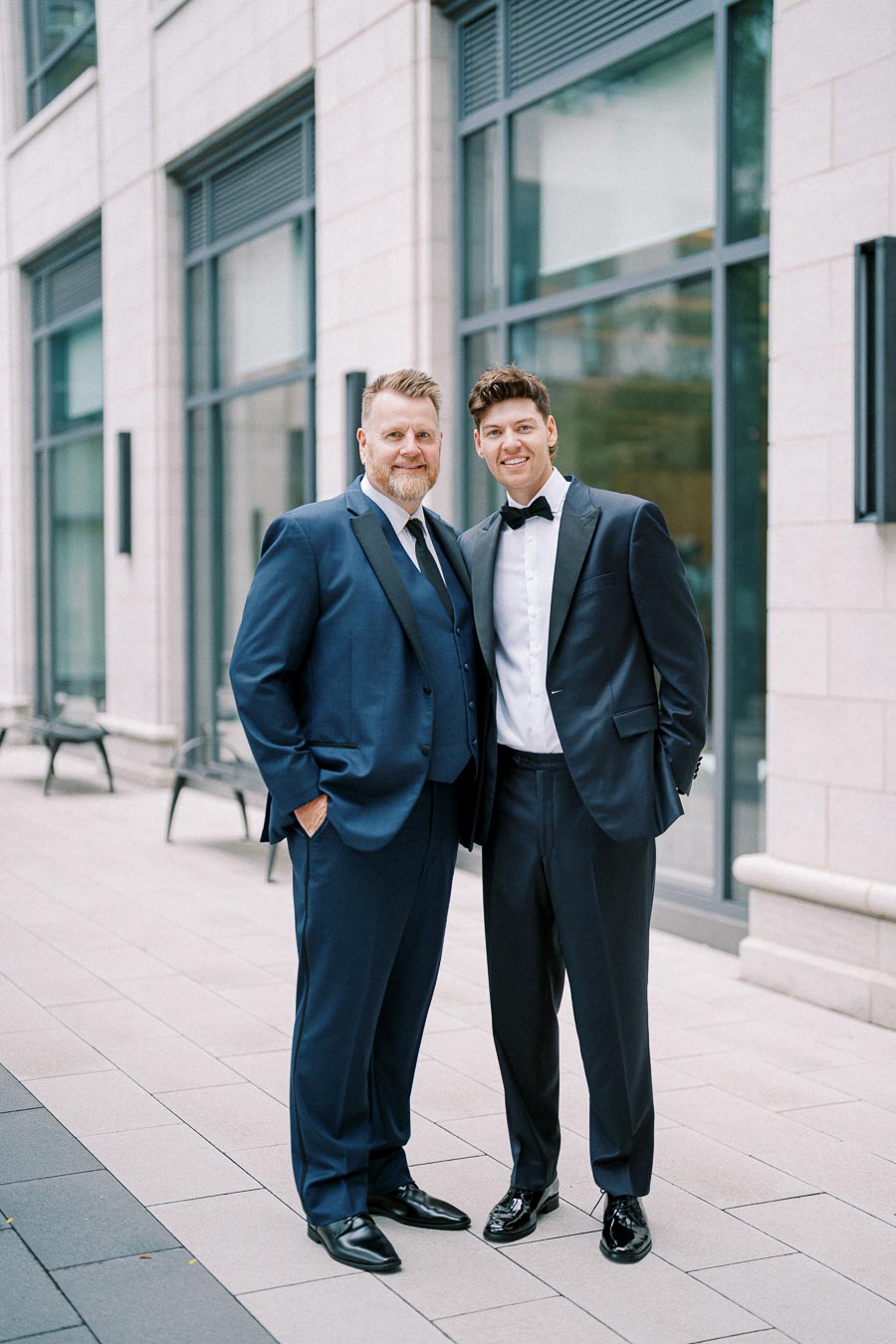 Two men in formal suits standing outside a modern building with large windows.