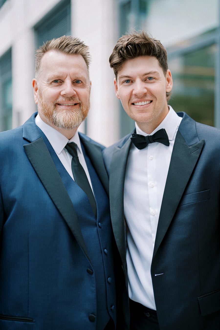 Two men in formal suits smiling at the camera, standing outdoors in a business district setting.