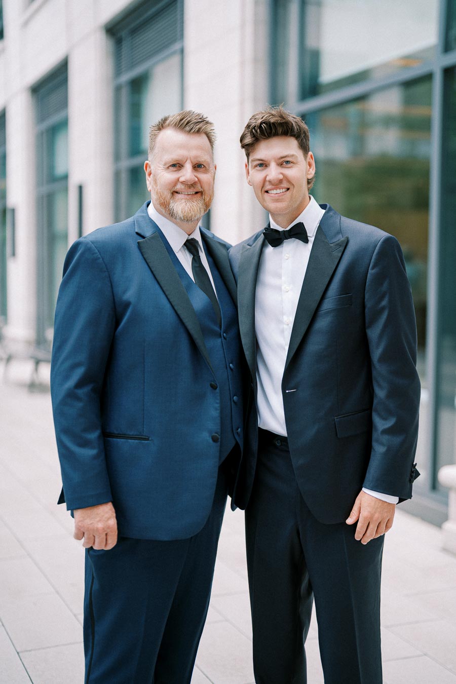 Two men in formal navy blue suits standing outside a modern building.