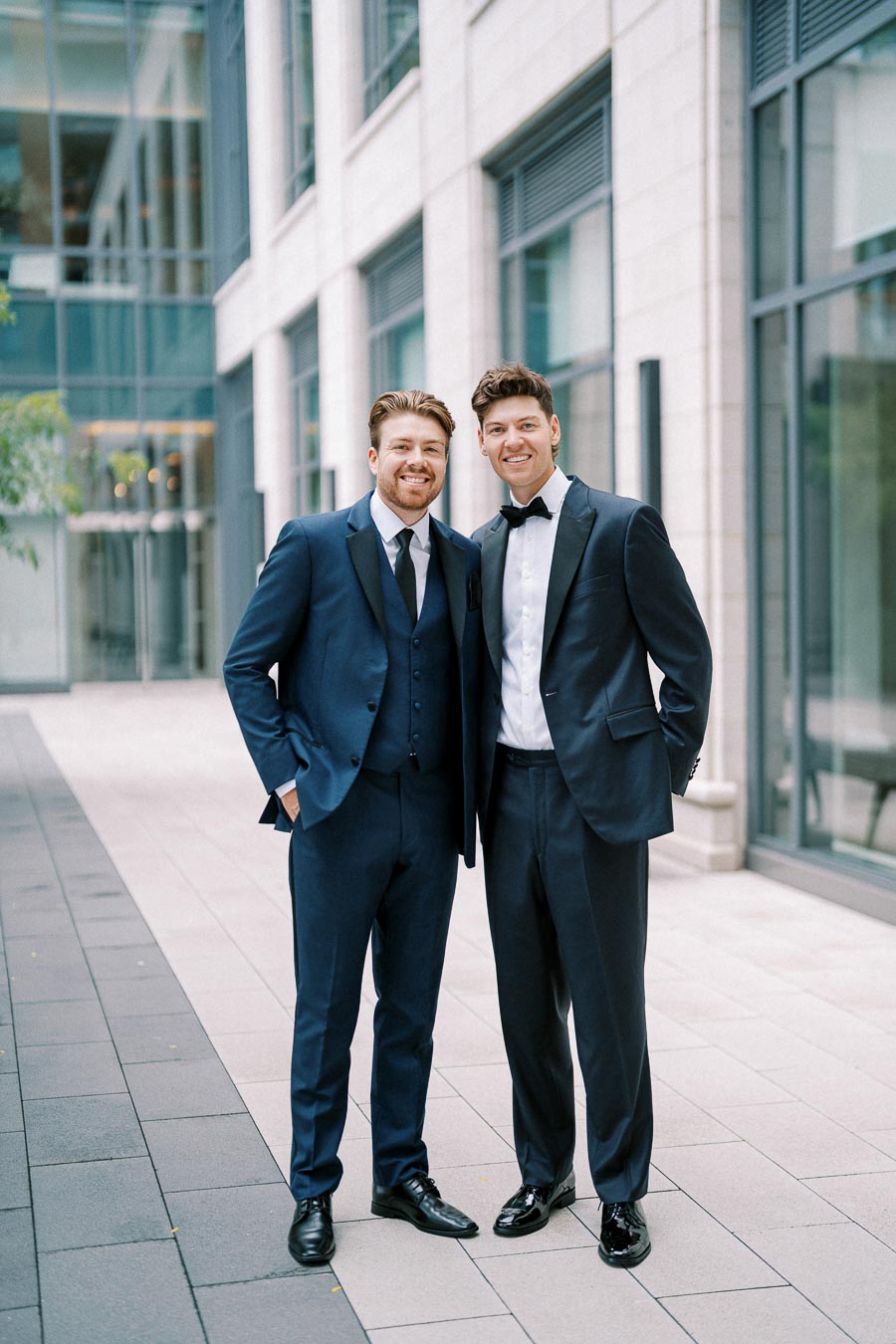 Two men dressed in formal suits pose confidently in an urban setting with modern architecture.