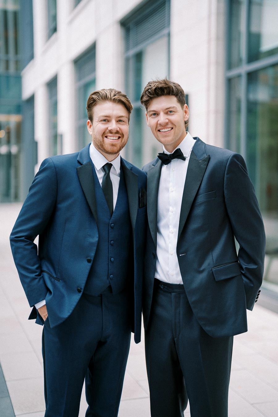 Two smiling men in formal suits standing outdoors in an urban setting, one wearing a three-piece suit and tie, and the other in a tuxedo with a bow tie, both with hands in their pockets.
