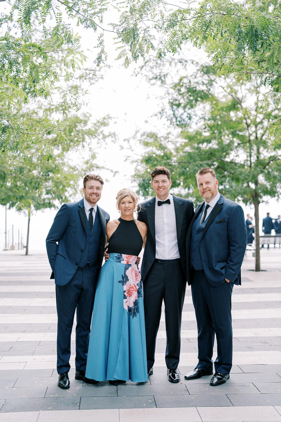A group of four well-dressed people posing outdoors under leafy trees, with the men in blue suits and a woman in a black top and blue floral skirt, showcasing elegant fashion at a formal event.