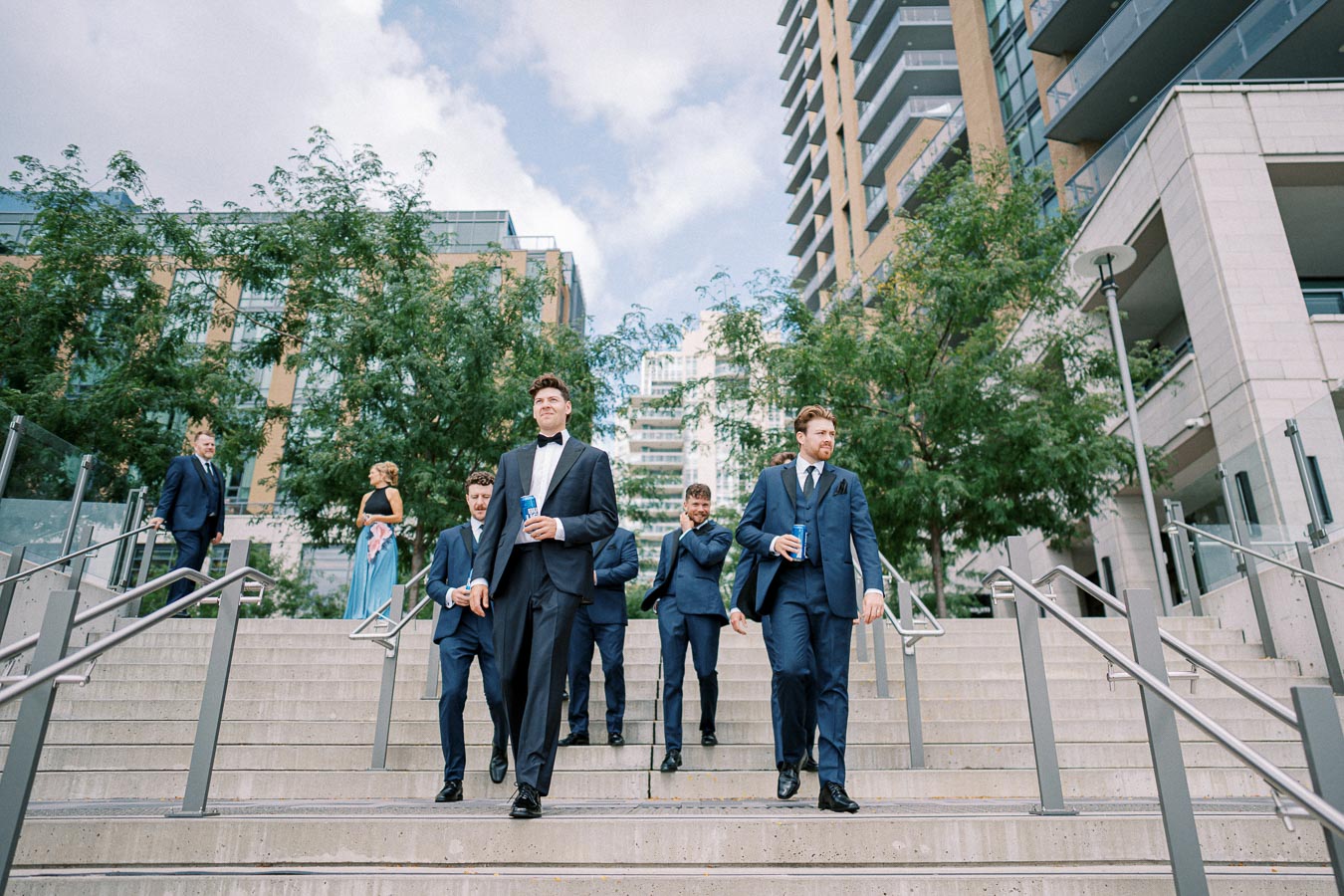 Group of men in suits, holding drinks, walk down a set of stairs outdoors in a modern urban setting.
