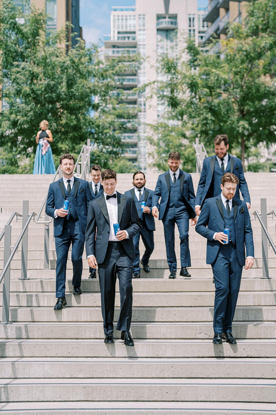 Groomsmen in blue suits descend concrete stairs outdoors, holding beverage cans, with urban buildings and greenery in the background on a sunny day.