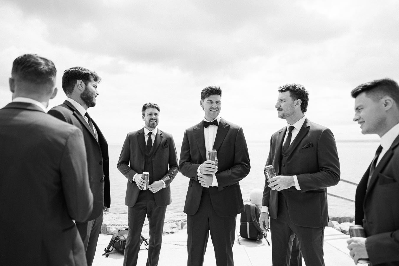 Group of men in formal suits enjoying drinks by the waterfront, engaged in conversation, perfect for a stylish wedding or sophisticated outdoor event setting.