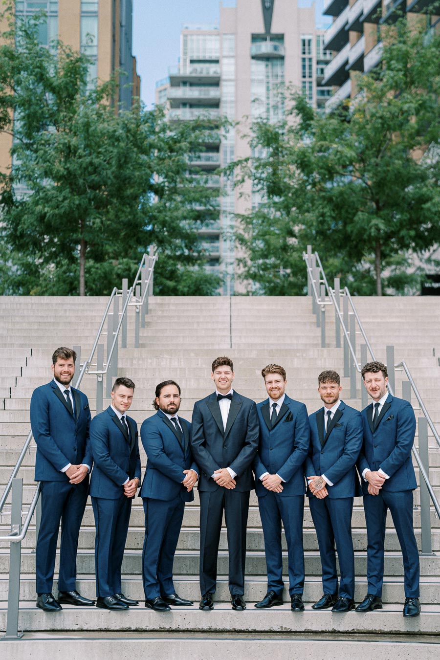 A group of groomsmen in matching navy suits and a groom in a tuxedo pose on outdoor stairs with urban buildings and greenery in the background, capturing a stylish and modern wedding setting.
