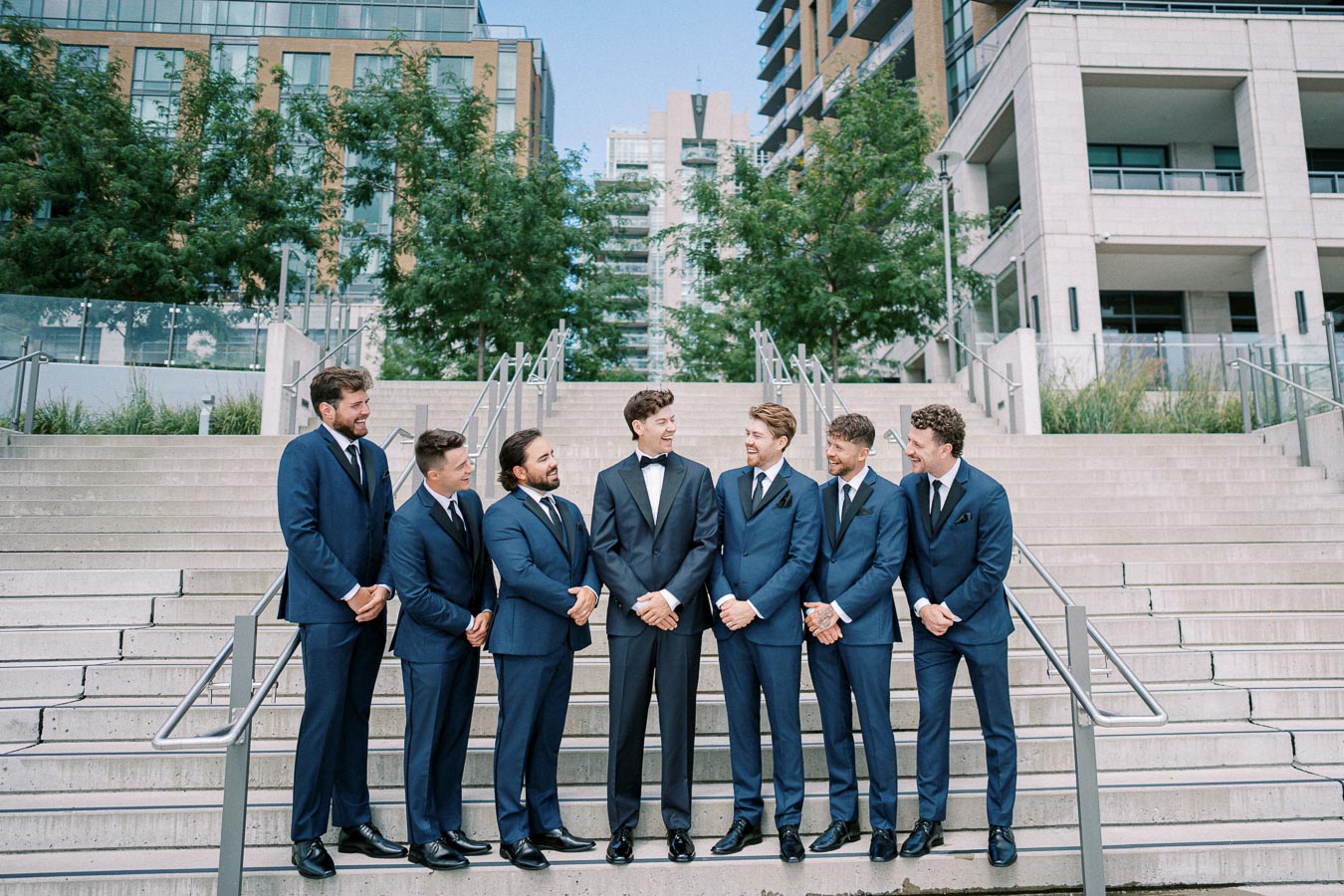 A group of groomsmen in matching blue suits and ties surround the groom, who is wearing a classic black tuxedo, posing on outdoor steps in an urban setting with modern buildings and trees in the background.