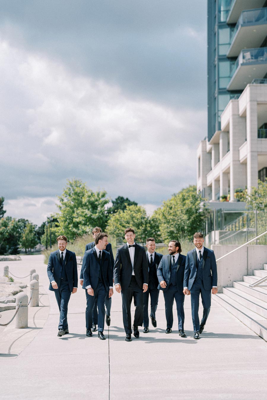 Group of groomsmen in navy suits and a groom in a tuxedo walking outside near a modern building on a cloudy day, capturing a joyful wedding moment