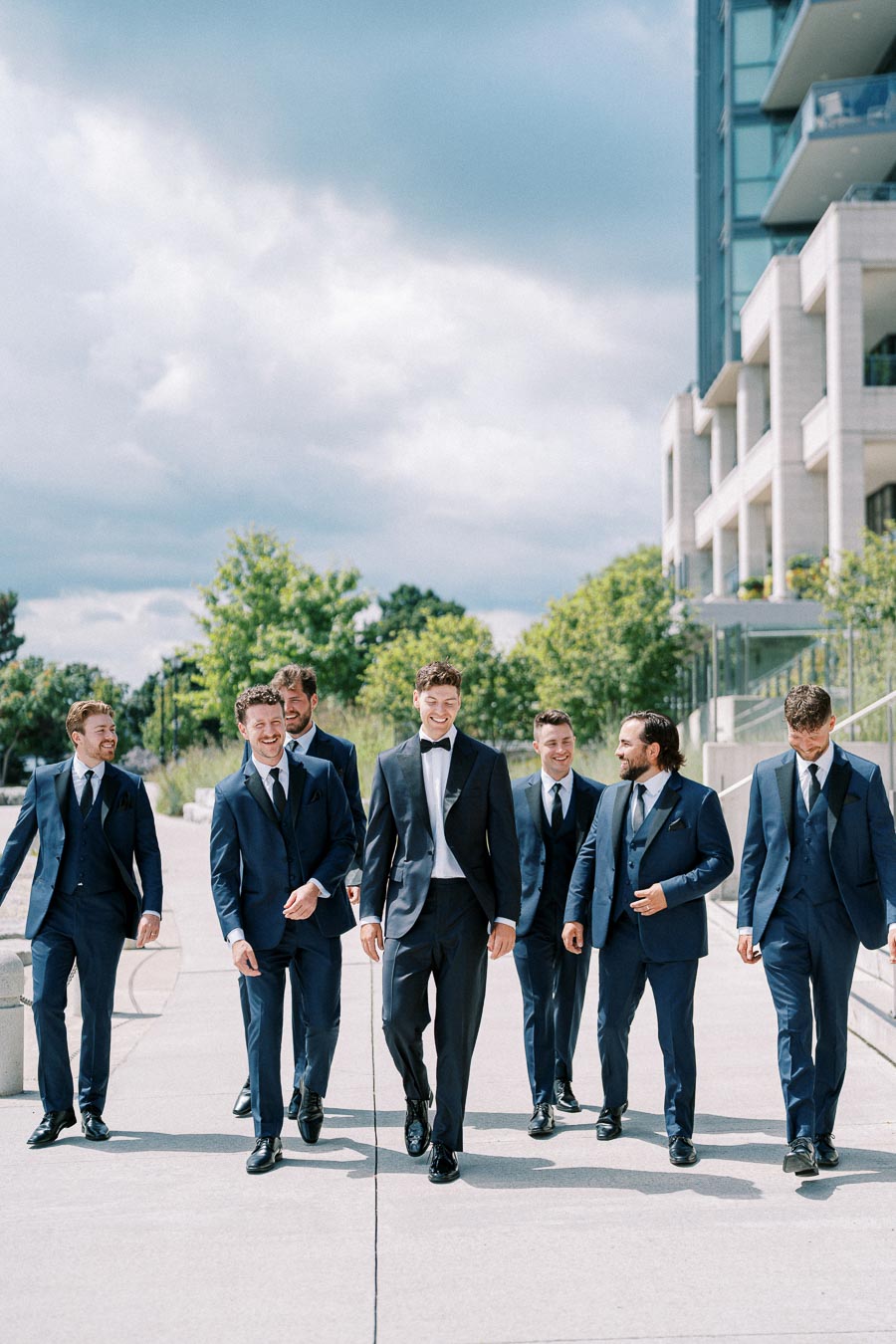 Group of seven men in matching blue suits walking together outdoors near a modern building, under a cloudy sky.