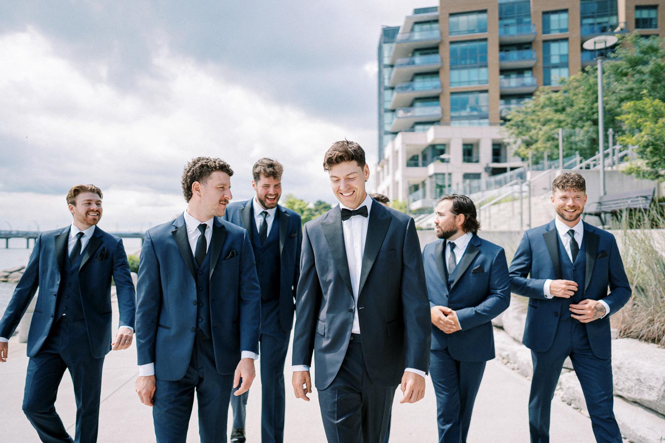 Groom and groomsmen walking in a modern cityscape, wearing matching navy blue suits and smiling, with a background of urban architecture and a cloudy sky.