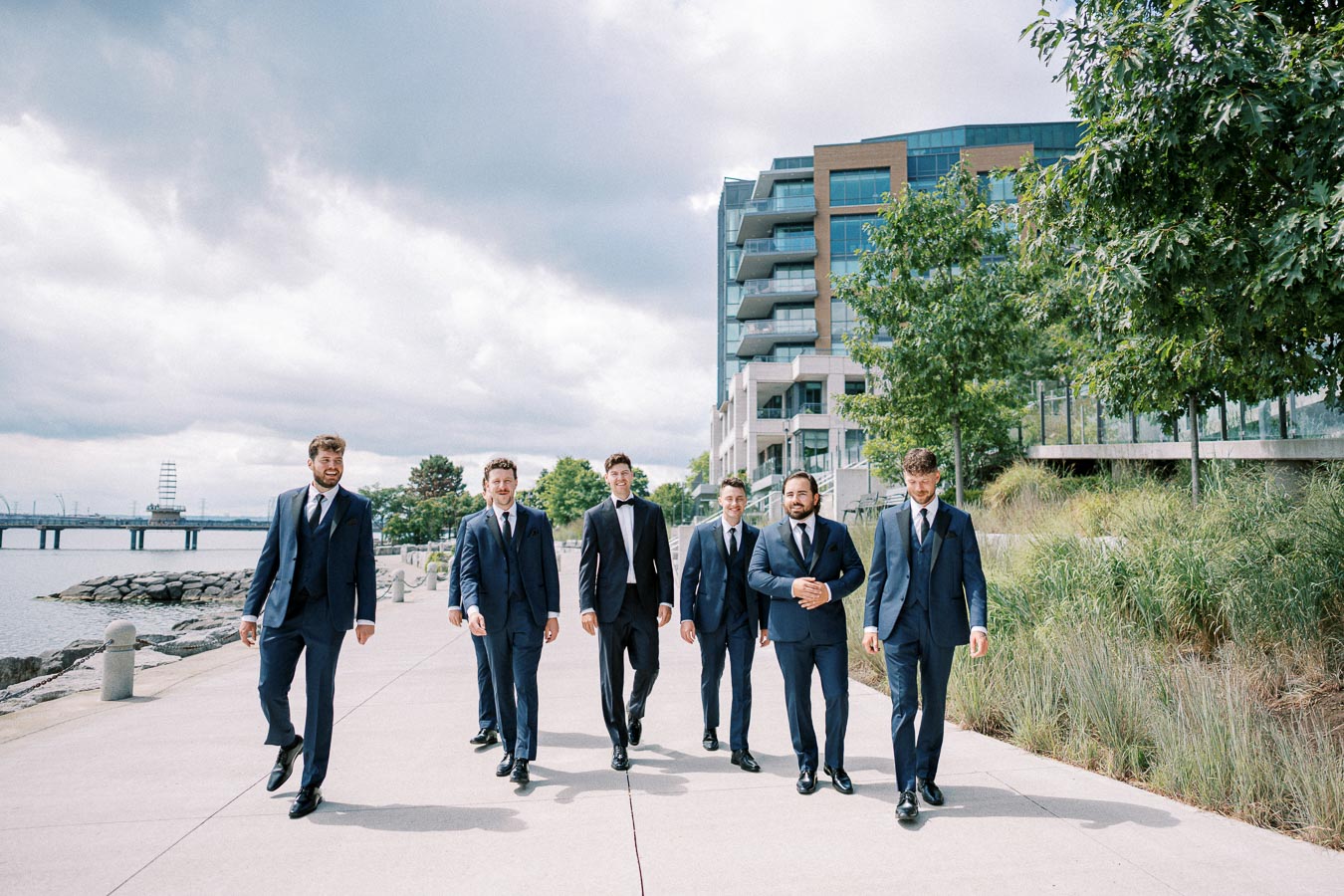 Groomsmen in matching blue suits walking along a waterfront pathway near modern buildings and greenery, with an overcast sky in the background.