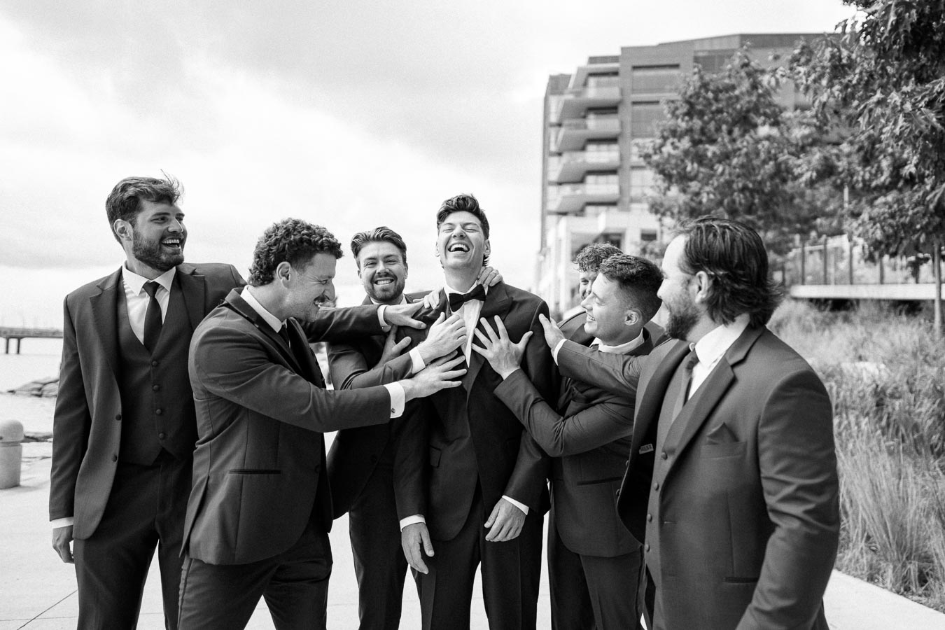 Group of groomsmen in suits laughing and playfully adjusting the groom's attire outdoors, captured in black and white.
