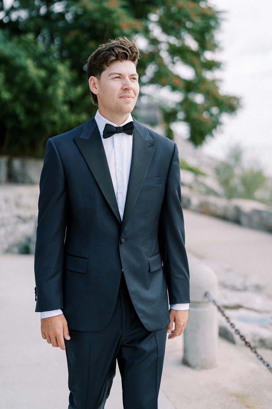 A man in a dark suit and bow tie standing outdoors on a path, with greenery in the background, exuding a formal and elegant appearance.