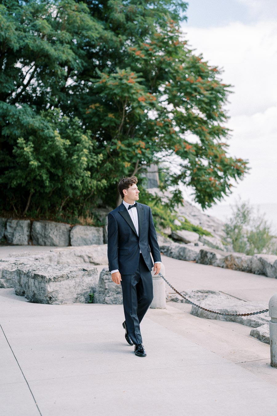 A man in a formal black tuxedo walks confidently along a paved path, surrounded by lush greenery and large rocks. The setting appears to be a peaceful outdoor location under a partly cloudy sky.