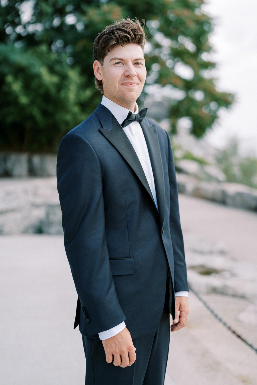 A man in a formal black tuxedo with a bow tie smiling outdoors, surrounded by greenery and stone pathways, exuding elegance and style.