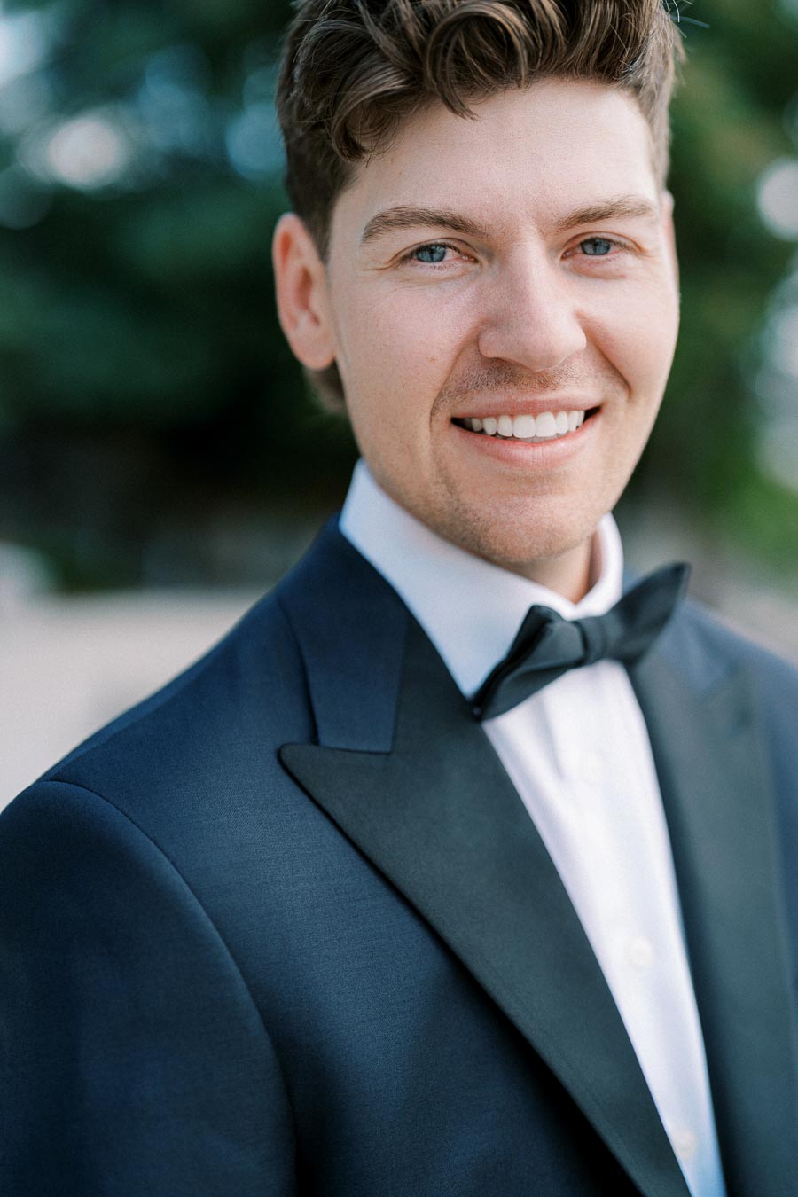 Man smiling in a tuxedo with a bow tie, outdoor background