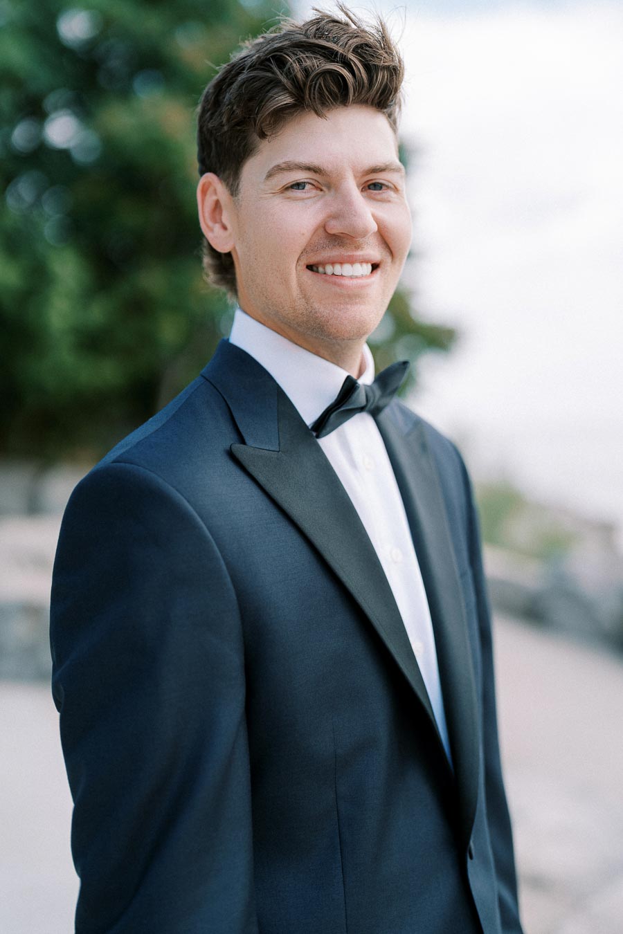 Smiling man in a tuxedo standing outdoors with trees in the background